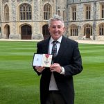 Jonathan Davies standing on the grounds of Windsor Castle holding his CBE medal and smiling.