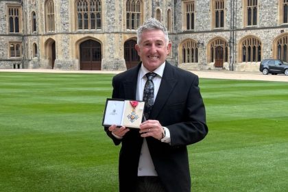Jonathan Davies standing on the grounds of Windsor Castle holding his CBE medal and smiling.