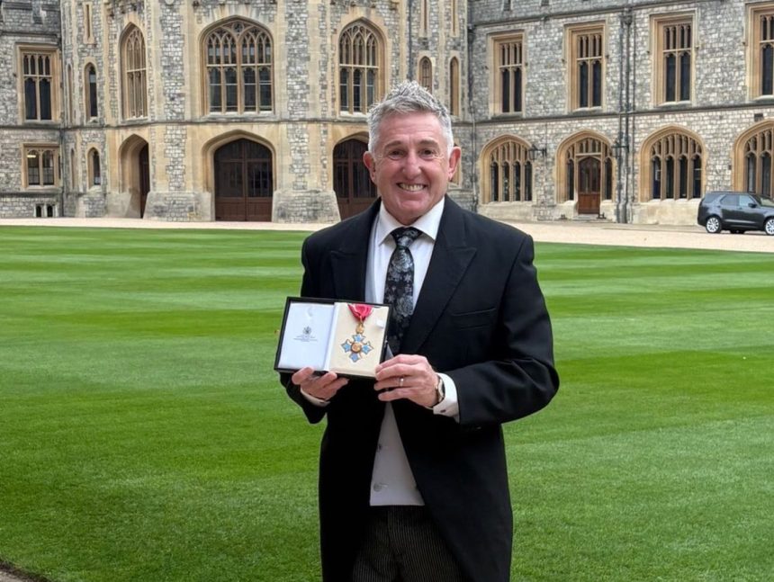 Jonathan Davies standing on the grounds of Windsor Castle holding his CBE medal and smiling.