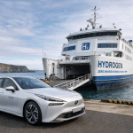 A white hydrogen-powered ferry docked at a coastal port with its vehicle ramp open, while a silver hydrogen car is parked in the foreground; cliffs and sea are visible in the background.