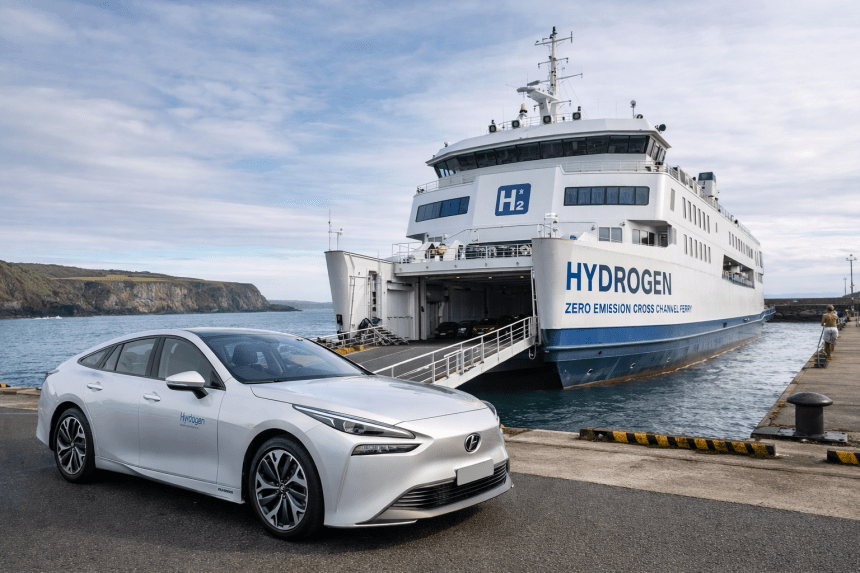 A white hydrogen-powered ferry docked at a coastal port with its vehicle ramp open, while a silver hydrogen car is parked in the foreground; cliffs and sea are visible in the background.