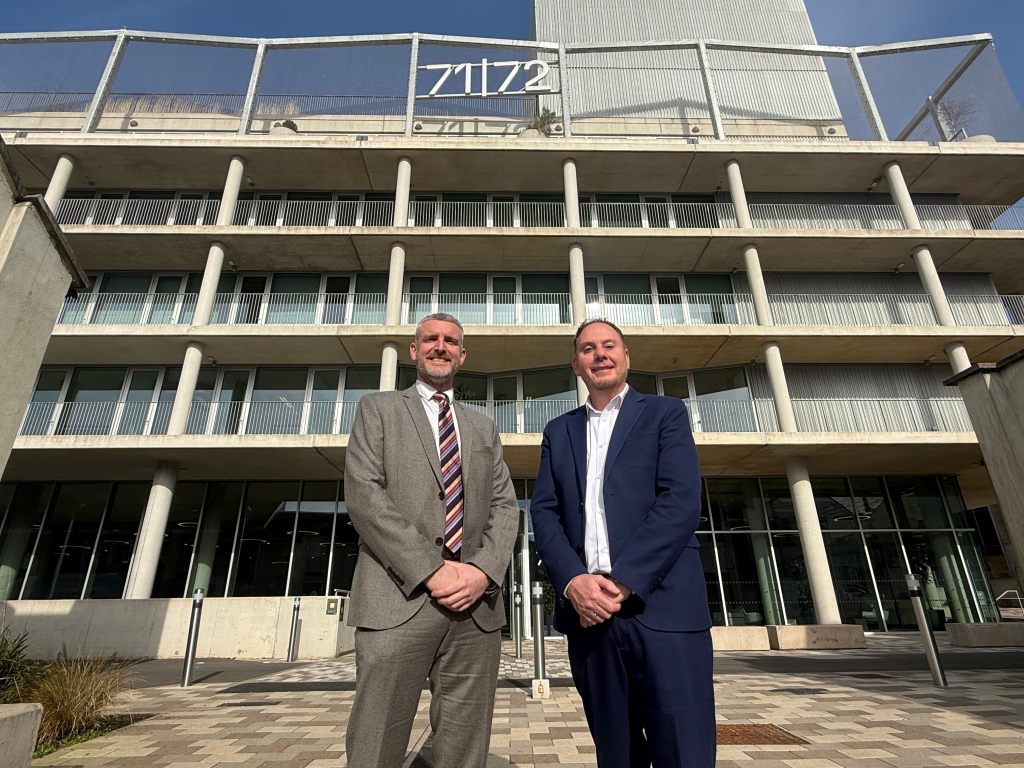 Two men in business attire standing outside a modern, multi-story building with a distinctive architectural design, featuring large windows and concrete balconies. The building has '71/72' prominently displayed at the top.