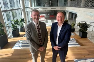 Two men in business attire standing inside a modern, spacious building with large windows, wooden flooring, and contemporary seating areas. One man is gesturing towards the other.