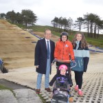 Four people posing for a photo on a dry ski slope. A man in a suit stands next to a woman in a ski jacket who is holding the handles of an adaptive sit-ski, in which a small child in a pink helmet is sitting. Another woman stands next to them.