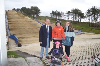 Four people posing for a photo on a dry ski slope. A man in a suit stands next to a woman in a ski jacket who is holding the handles of an adaptive sit-ski, in which a small child in a pink helmet is sitting. Another woman stands next to them.