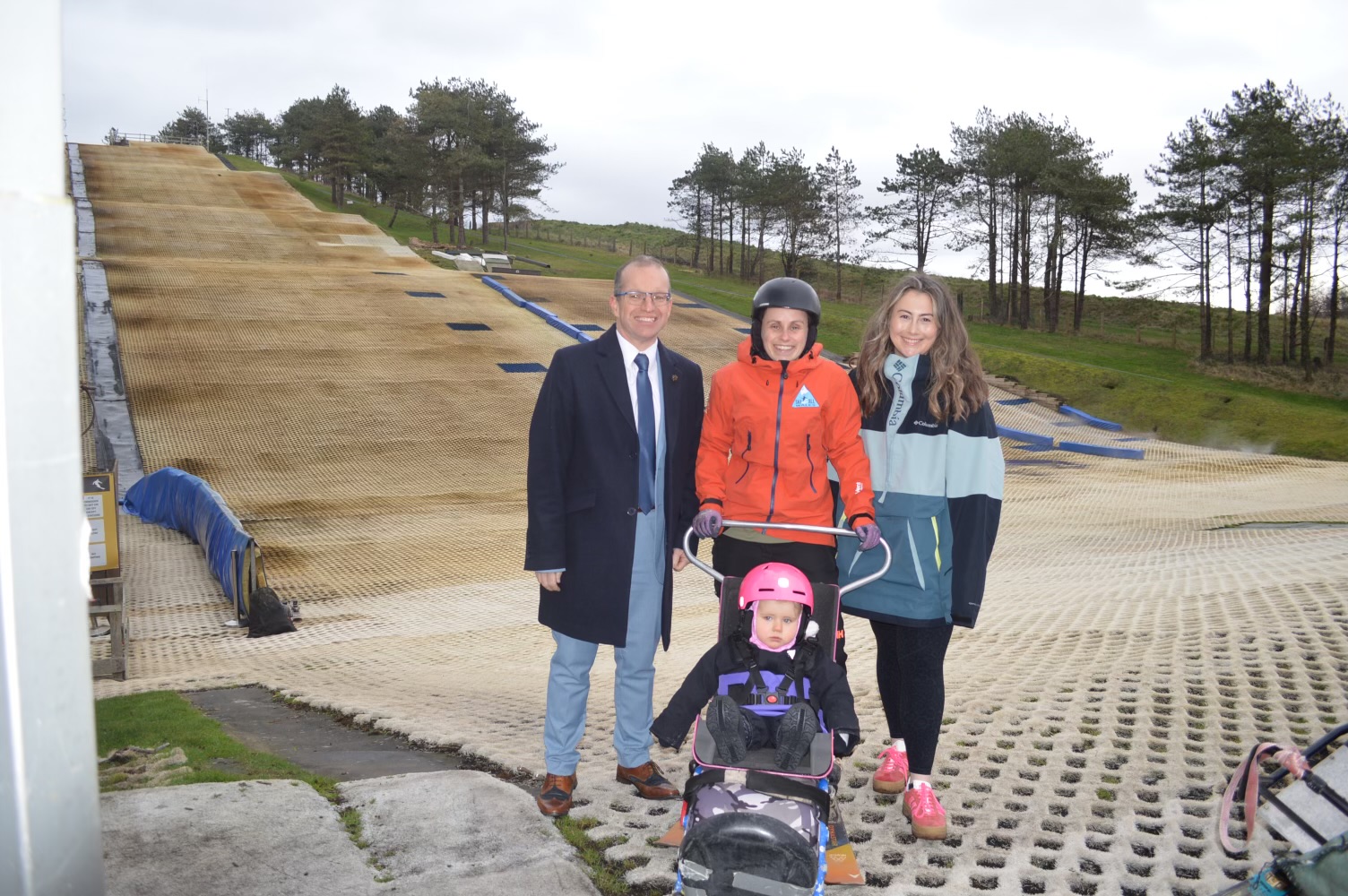 Four people posing for a photo on a dry ski slope. A man in a suit stands next to a woman in a ski jacket who is holding the handles of an adaptive sit-ski, in which a small child in a pink helmet is sitting. Another woman stands next to them.