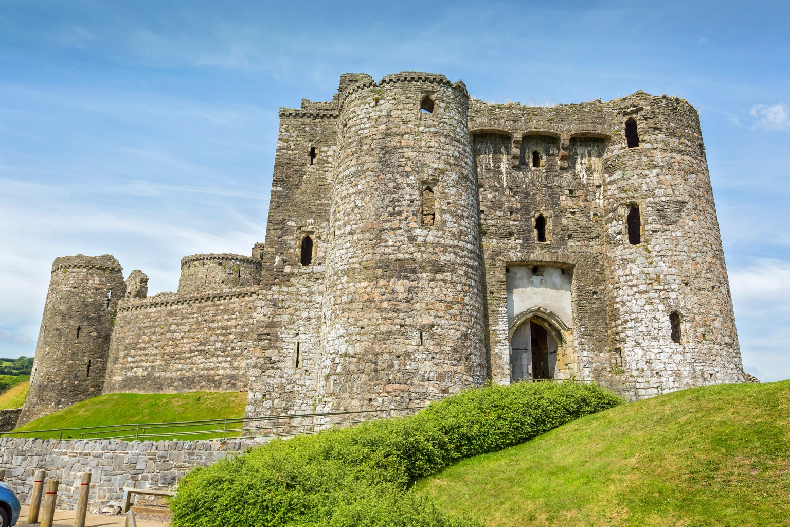 A large, medieval stone castle with tall cylindrical towers and a fortified gatehouse under a blue sky. The castle is built on a grassy mound with a stone wall and green bushes in the foreground.