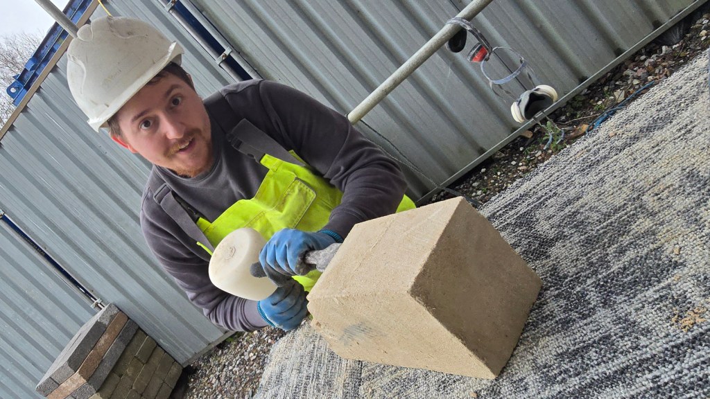 A stonemason in a hard hat and hi-vis vest chisels a pale stone block at the Laboratory Building restoration site.