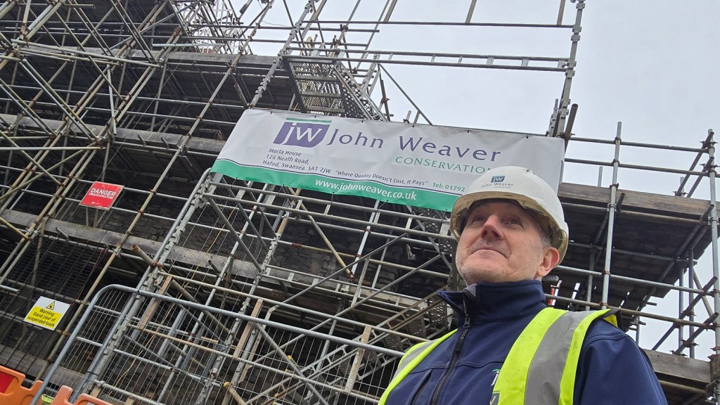 Simon Williams, wearing a hi-vis jacket and hard hat, stands in front of the scaffolded Laboratory Building with a John Weaver Conservation site banner visible behind him.