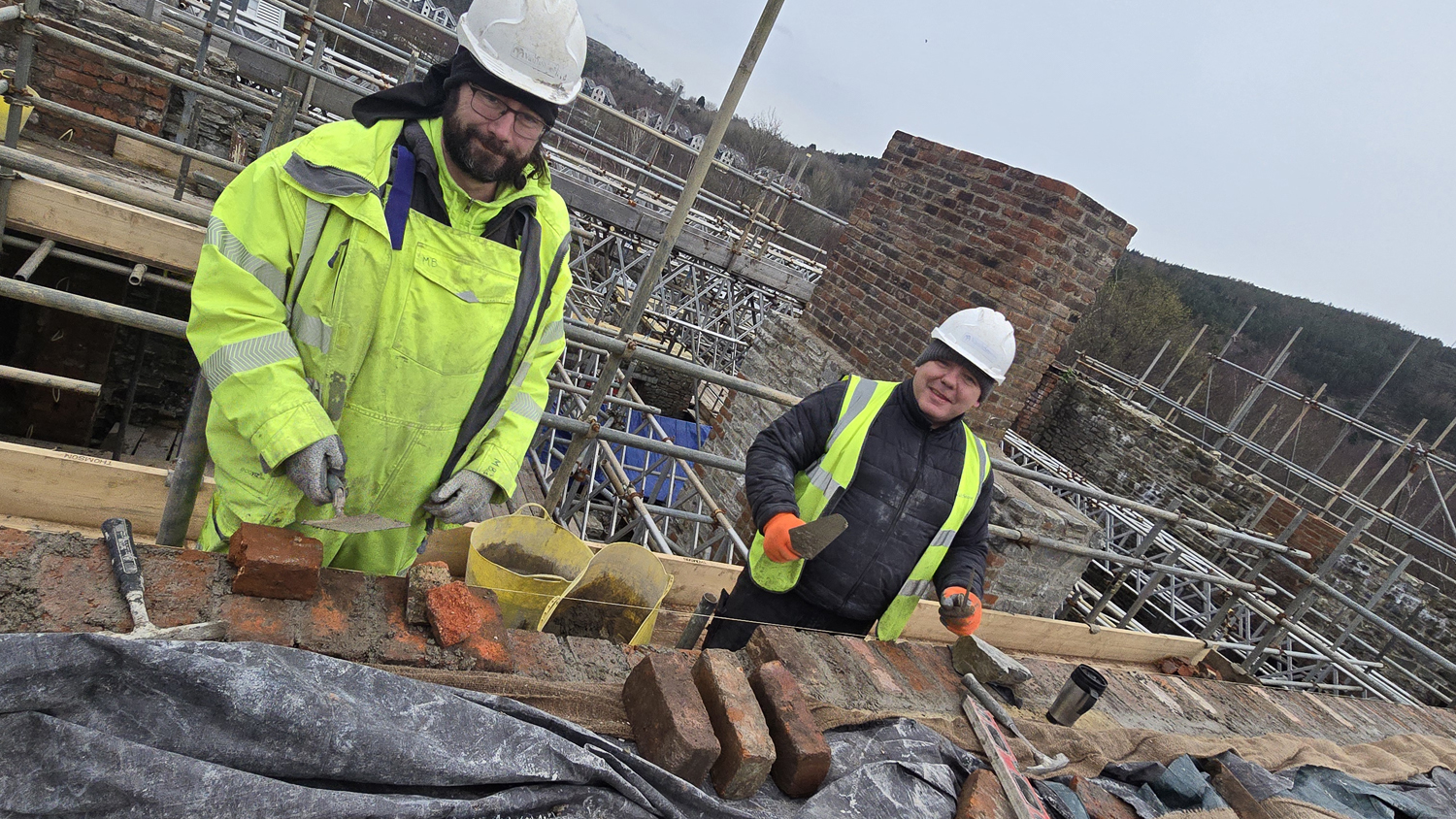 Two stonemasons in hi-vis jackets and hard hats lay bricks on scaffolding during restoration work on the Laboratory Building at the Hafod-Morfa Copperworks site in Swansea.