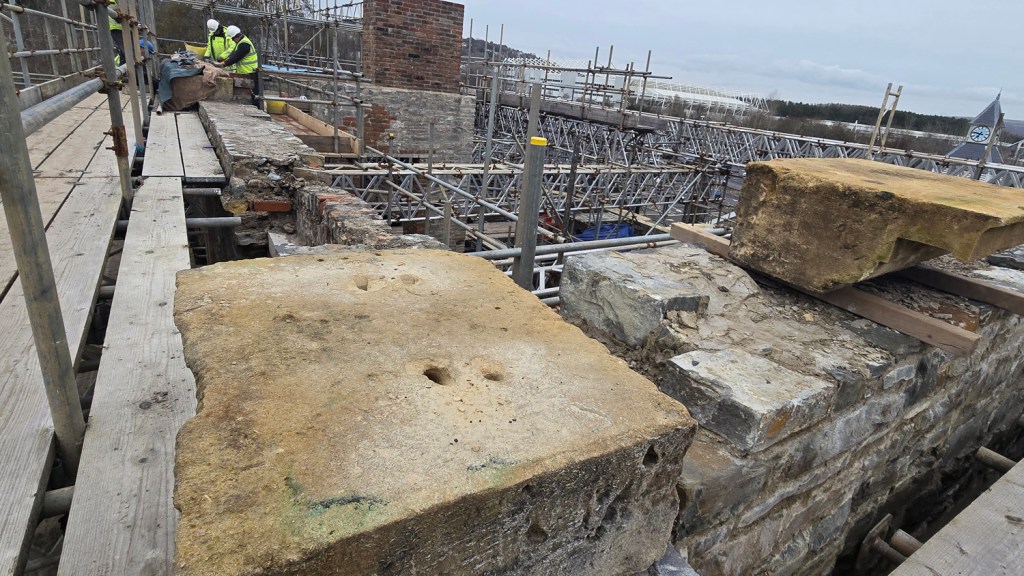 Large pale stone blocks sit on scaffolding boards at the Laboratory Building restoration site, with workers visible in the background and the Swansea Arena clock tower in the distance.