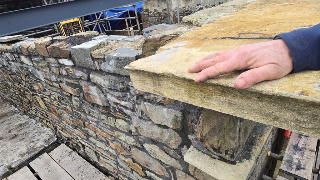 a hand rests on a newly installed stone cornice course above a repaired rubble stone wall.