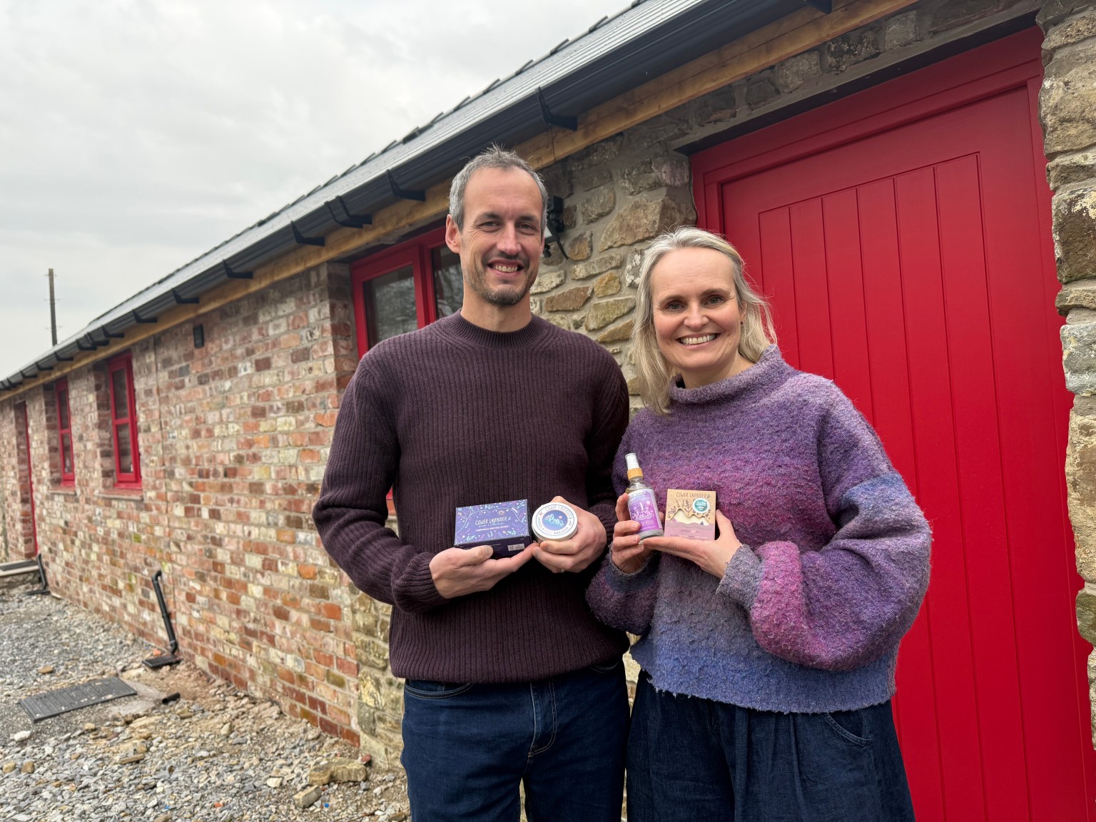 A man and a woman stand smiling outside a refurbished red-brick barn with a bright red door, holding up small jars and bottles of their lavender products.