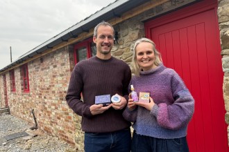 A man and a woman stand smiling outside a refurbished red-brick barn with a bright red door, holding up small jars and bottles of their lavender products.
