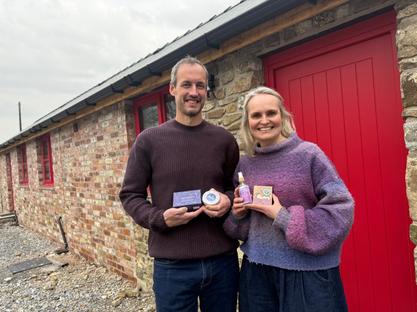 A man and a woman stand smiling outside a refurbished red-brick barn with a bright red door, holding up small jars and bottles of their lavender products.