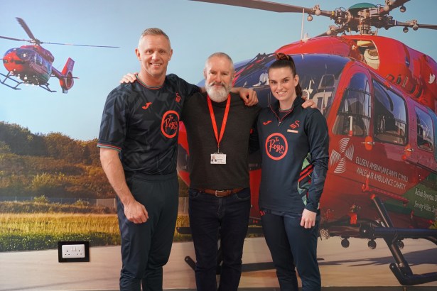 Lee Trundle, Christian Sullivan and Sammy Wynne stand together for a photo in front of a large wall graphic of a Wales Air Ambulance helicopter.