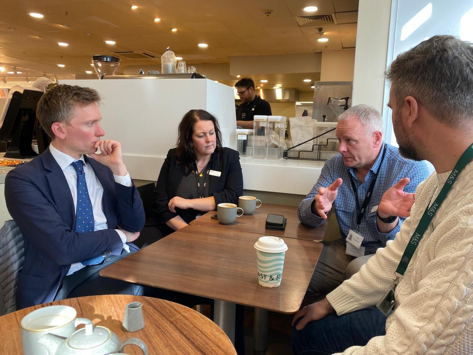 Swansea West MP Torsten Bell, wearing a suit and tie, sits at a cafe table with three other people, including M&S representatives, during an urgent meeting to discuss the closure of the city's Marks and Spencer store.