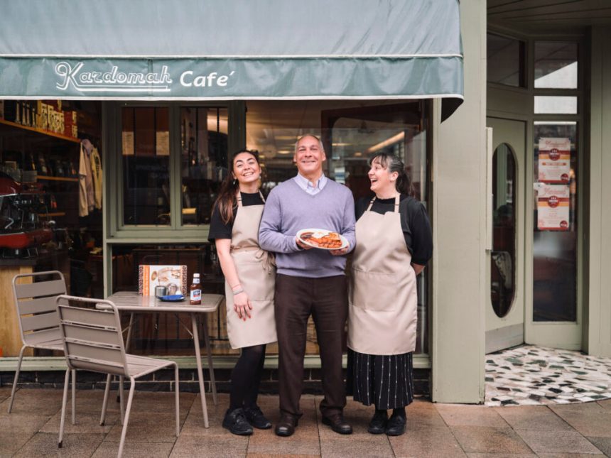 Three people stand outside Kardomah Cafe in Swansea, with owner Marcus Luporini holding a plate of breakfast food alongside two staff members wearing aprons at the café entrance.