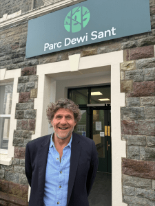 A man with curly dark hair, wearing a blue shirt and dark jacket, stands smiling in front of a stone building with a sign that reads “Parc Dewi Sant”.