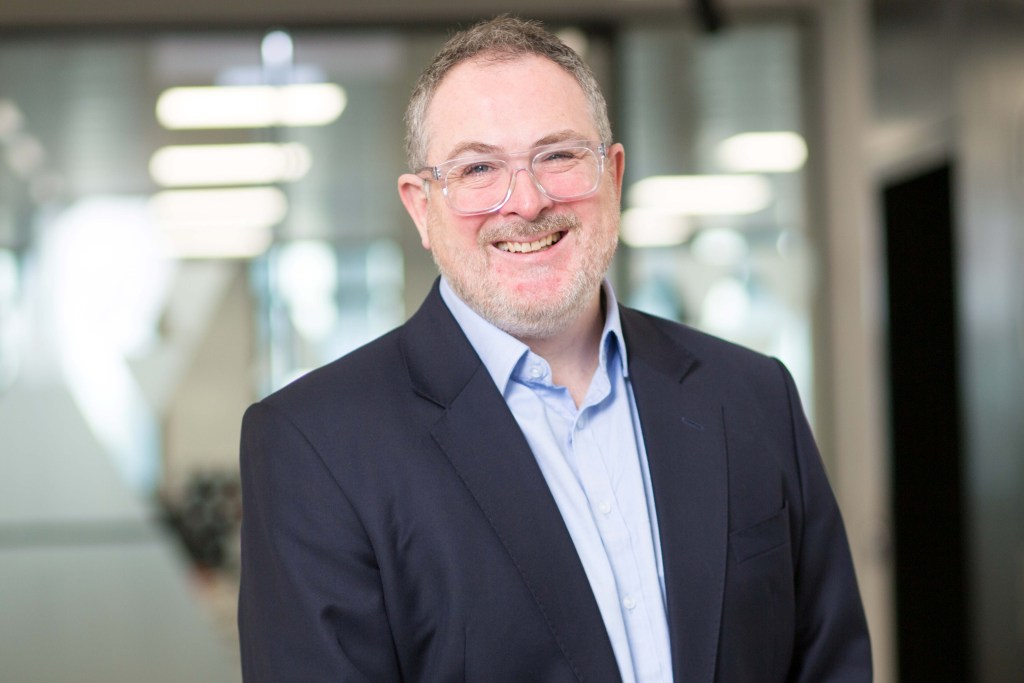 A professional headshot of Fraser Campbell, a smiling man with glasses and a beard, wearing a navy blazer and light blue shirt, in a modern office setting.