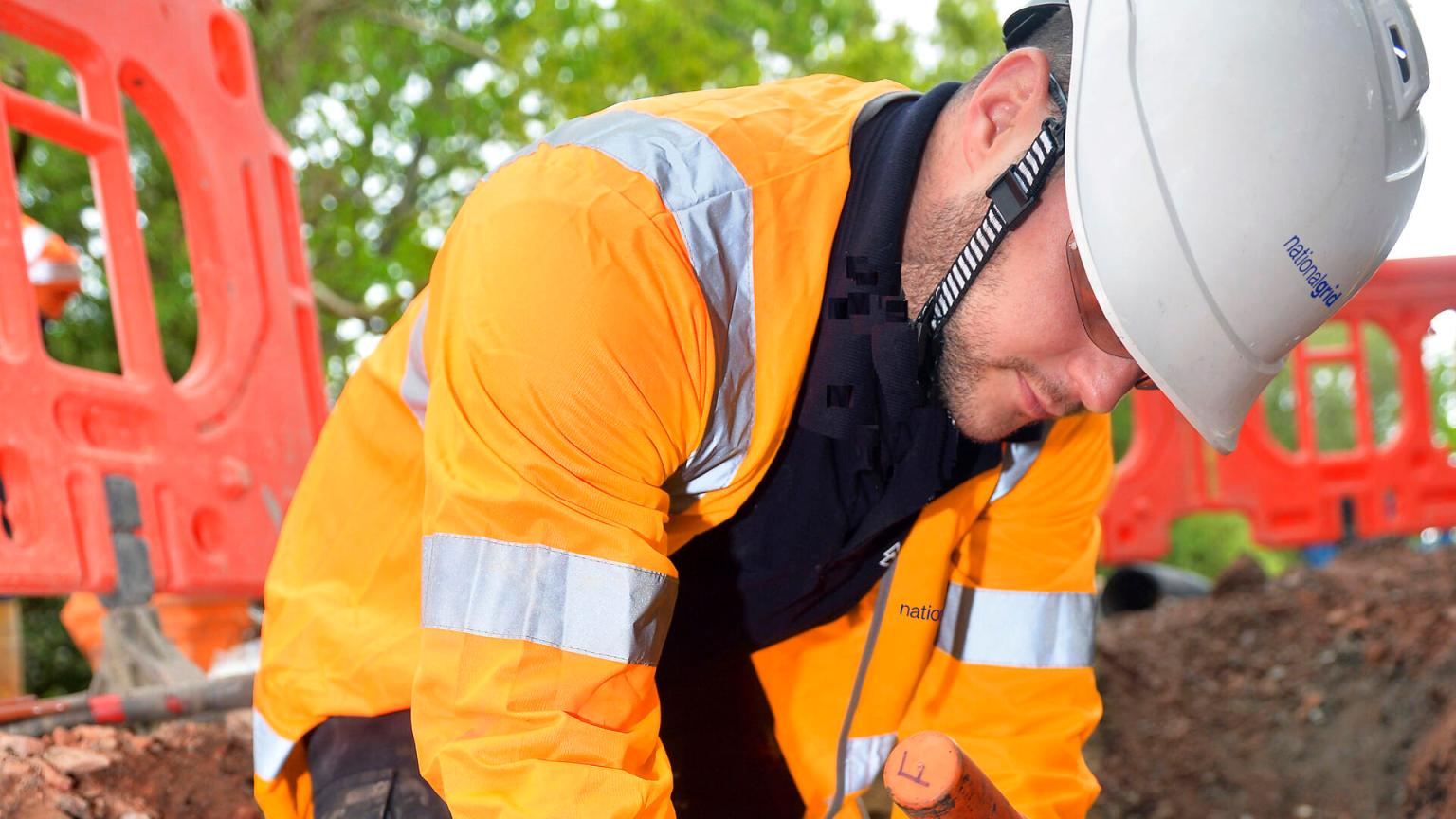 A National Grid worker in an orange high-visibility jacket and white hard hat bends over to work on underground cables at a roadside excavation site, with red safety barriers in the background.