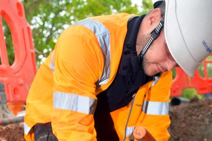A National Grid worker in an orange high-visibility jacket and white hard hat bends over to work on underground cables at a roadside excavation site, with red safety barriers in the background.