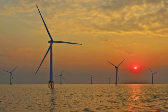 Several large offshore wind turbines stand in the sea against a golden sunset. The sun is a bright red orb low on the horizon, and its light reflects on the calm water.