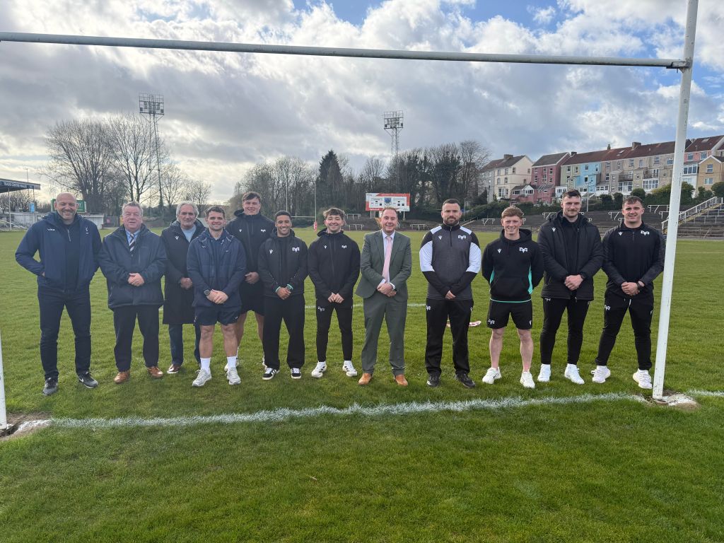 Group of Ospreys players and officials standing on rugby pitch at St Helens Swansea after partnership announcement