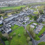 An aerial photograph showing a large, sprawling complex of historic stone buildings with grey slate roofs, surrounded by green lawns, trees, and roads with car parks.