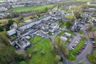 An aerial photograph showing a large, sprawling complex of historic stone buildings with grey slate roofs, surrounded by green lawns, trees, and roads with car parks.