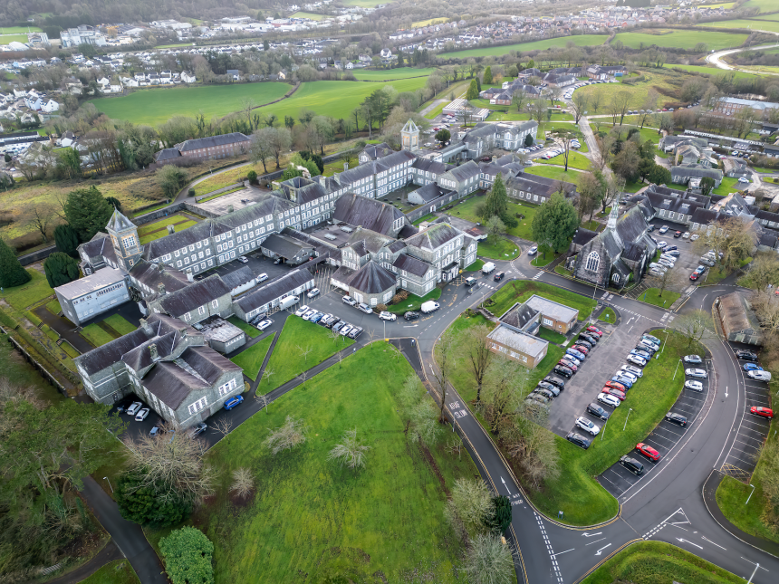An aerial photograph showing a large, sprawling complex of historic stone buildings with grey slate roofs, surrounded by green lawns, trees, and roads with car parks.