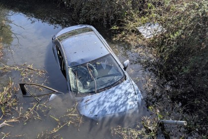 A close-up, landscape crop showing the front of a silver car submerged in water with surrounding reeds and vegetation, taken during an emergency response.