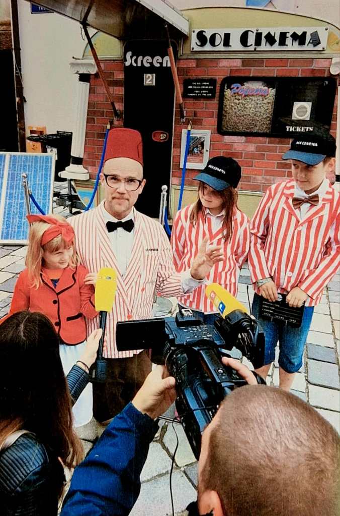 A small pop-up cinema setup with people in vintage-style outfits speaking to media in front of a “Sol Cinema” sign.