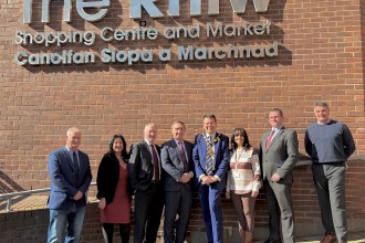 A group of council leaders and officials standing outside The Rhiw Shopping Centre sign in Bridgend on a sunny day.