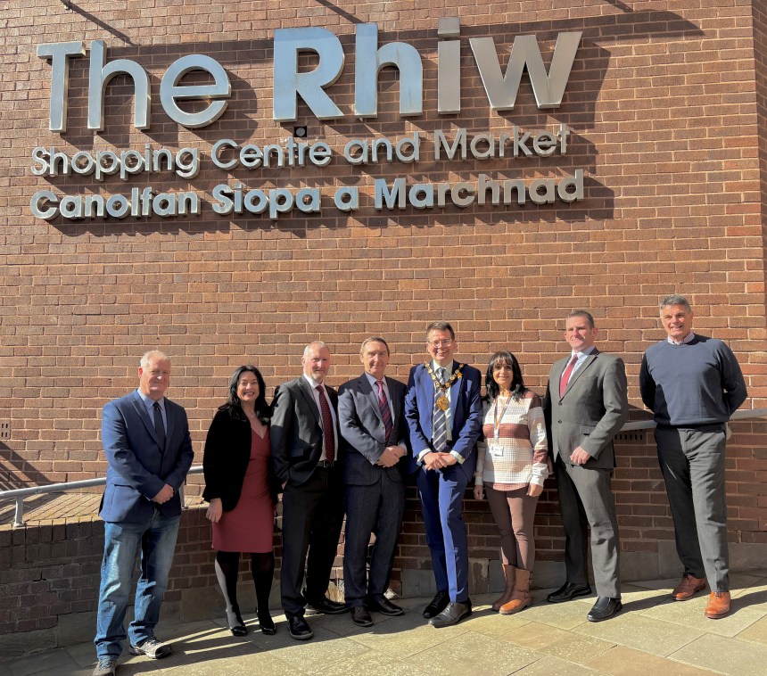 A group of council leaders and officials standing outside The Rhiw Shopping Centre sign in Bridgend on a sunny day.