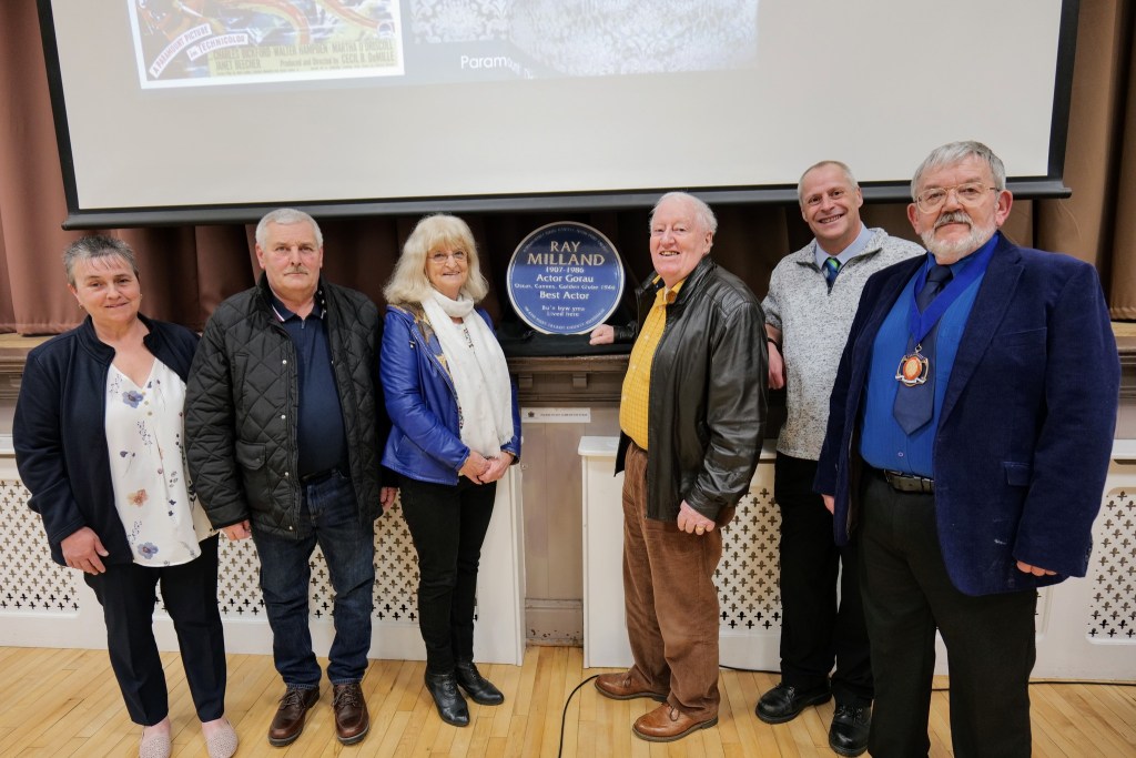 A group of people standing in a hall behind a blue plaque for Ray Milland, which is resting on a mantelpiece. They are smiling and celebrating the installation.