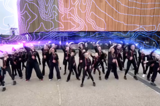 A large group of young dancers dressed in black perform a choreographed routine outside the Swansea Arena, with the building's illuminated LED display visible behind them.