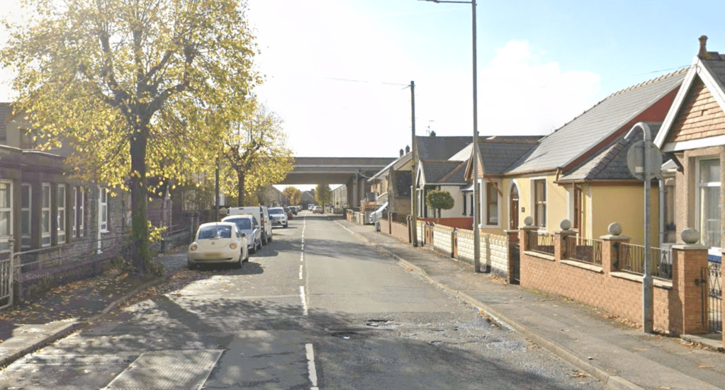 A residential street in Port Talbot lined with bungalows and parked cars, with a road bridge visible in the background and autumn trees to the left.