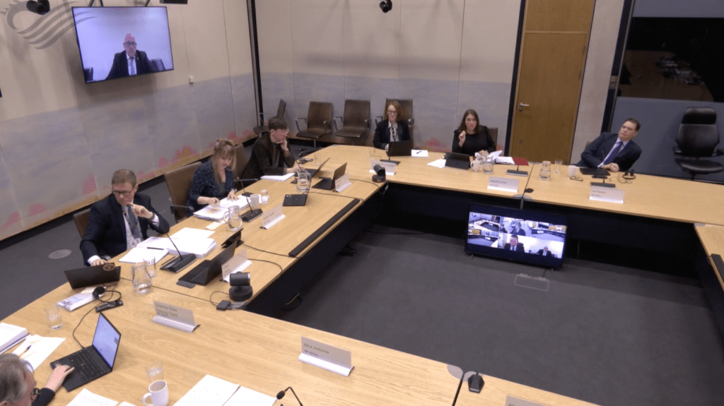 A wide-angle view of a Senedd committee room, with members seated around a large rectangular table. A screen on the wall shows WRU Chair Richard Collier-Keywood appearing via video link.