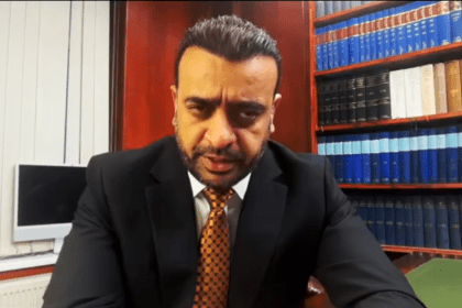A close-up screenshot of Ahsan Ullah, CEO of Ullcom Group, wearing a suit and patterned tie, speaking directly to the camera in front of a bookshelf filled with blue volumes.