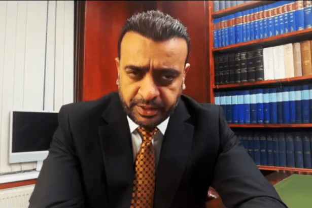 A close-up screenshot of Ahsan Ullah, CEO of Ullcom Group, wearing a suit and patterned tie, speaking directly to the camera in front of a bookshelf filled with blue volumes.
