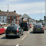 A street-level view of Fforestfach Cross in Swansea, showing multiple lanes of cars and vans queuing at a busy junction with traffic lights. Several red, black, and grey vehicles are visible in the foreground under a clear sky.