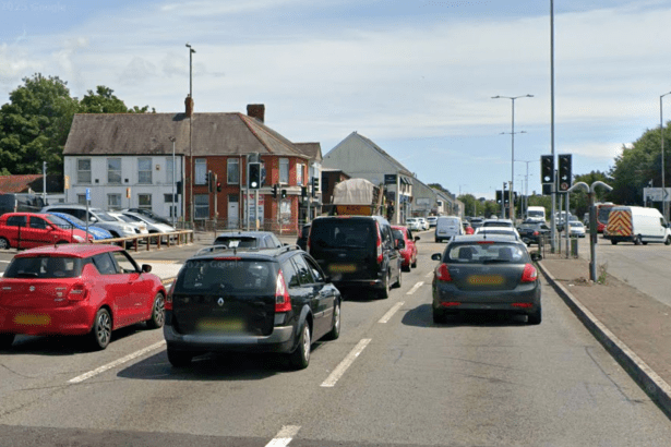 A street-level view of Fforestfach Cross in Swansea, showing multiple lanes of cars and vans queuing at a busy junction with traffic lights. Several red, black, and grey vehicles are visible in the foreground under a clear sky.
