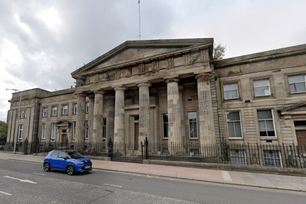 A street-level view of the historic High Court building in Glasgow, featuring large stone columns and a classical facade. A blue car is seen driving past on the road in the foreground under an overcast sky.