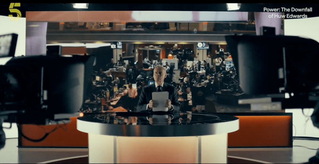 A wide shot of Martin Clunes as Huw Edwards sitting behind a sleek, circular news desk in a large, busy newsroom setting. Two studio cameras are visible in the foreground, framing the shot.