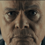 An extreme close-up of Martin Clunes' face, transformed with make-up and silver hair to resemble Huw Edwards. He has a stern, intense expression, looking directly into the camera.