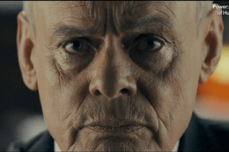 An extreme close-up of Martin Clunes' face, transformed with make-up and silver hair to resemble Huw Edwards. He has a stern, intense expression, looking directly into the camera.