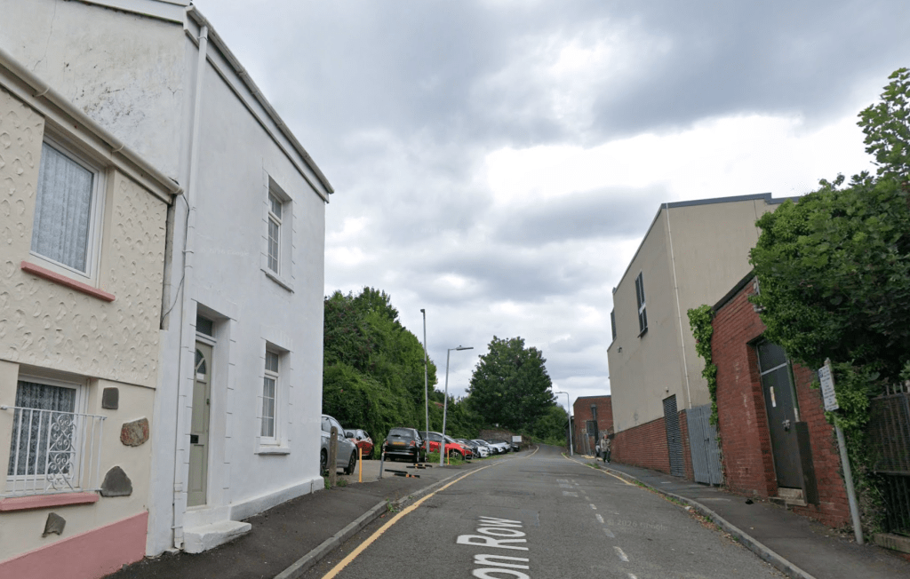 A street-level view looking down Clifton Row in Swansea. White residential houses are on the left, and the back of a large brick building is on the right. Several cars are parked in a small lot further down the narrow road under an overcast sky.