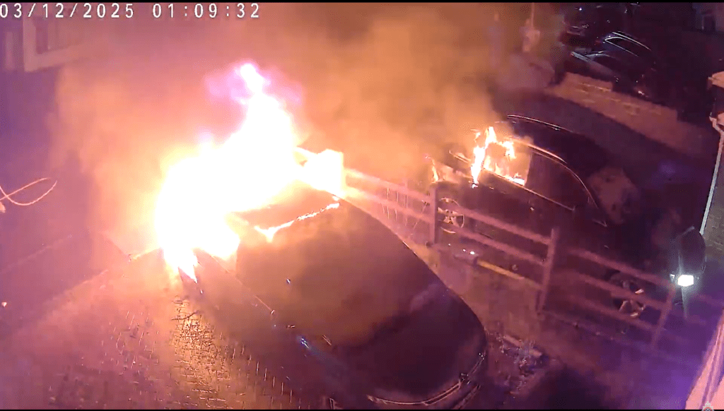 CCTV image showing a car on fire in a residential street in Brackla, Bridgend, with flames spreading to a nearby vehicle at night.