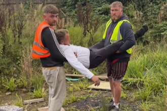 Two tradesmen in high-visibility vests are playfully carrying a third person in a white top, who is stretched out horizontally between them. They are outdoors in a grassy, somewhat overgrown area.